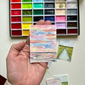 May include: A small watercolor painting of a sunset with pink, blue, and yellow hues. The painting is held in a hand, with a watercolor paint set in the background. Additional small paintings are also visible.
