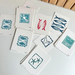 May include: A collection of small, square art prints featuring various sea-themed linocut designs. The prints showcase images of fish, a crab, a whale tail, starfish, and seagulls in shades of teal and red. Each print is on white paper.