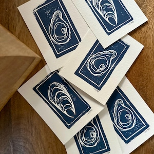 May include: A collection of handmade art prints featuring a navy blue oyster design on off-white paper. The prints are arranged on a wooden surface, showcasing the linocut style and the contrast between the dark blue and light paper.
