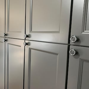 May include: Gray kitchen cabinets with decorative silver trim and round, crystal-studded knobs. The cabinets have a recessed panel design and are arranged in a row. The doors are closed, and the overall style is modern and sleek.
