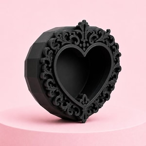 May include: A black heart-shaped decorative box with an ornate, scrollwork frame. The box has a faceted exterior and sits on a pink pedestal. The heart-shaped opening is visible in the center of the frame.