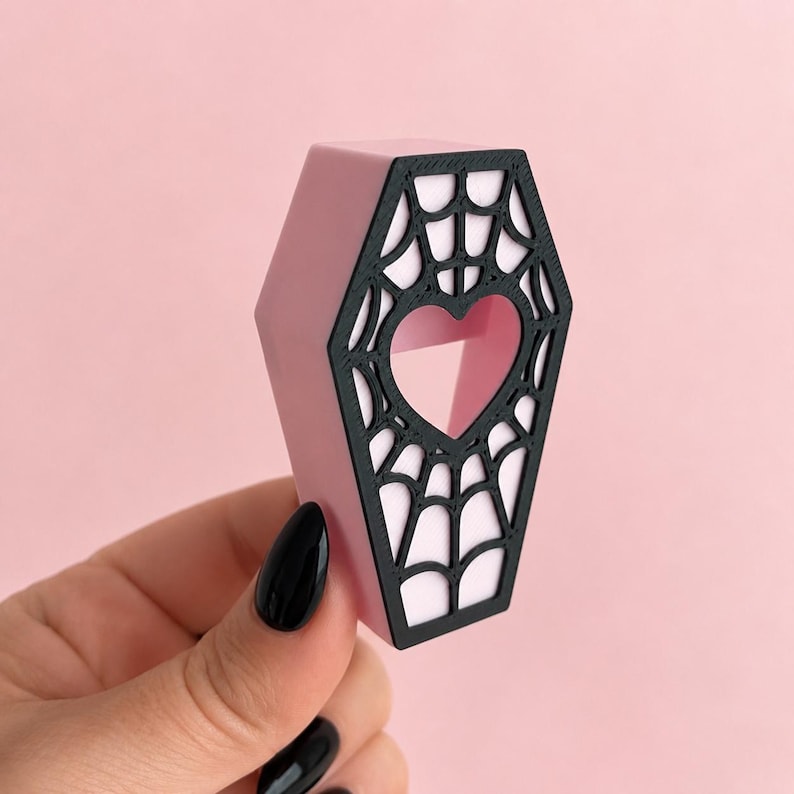 Hand holding a purple magnetic jumping spider coffin hide with black spiderweb detail on a pastel pink background.