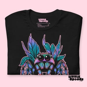 May include: Black t-shirt with a colorful illustration of a jumping spider with iridescent wings. The spider has large, round eyes and is surrounded by a floral design. The shirt has the text "LITTLE -SPOODS" on the front.