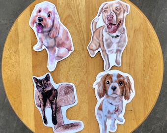 May include: Four pet stickers on a wooden surface. The stickers feature illustrations of a Cockapoo, a Pitbull, a cat, and a Cavalier King Charles Spaniel. The Cockapoo is light brown, the Pitbull is tan, the cat is black and brown, and the Cavalier is brown and white.