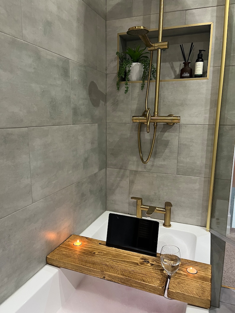May include: A wooden bath tray with a built-in phone or tablet holder, a glass of water, and two candles. The tray is placed on a white bathtub.