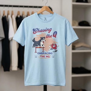 Könnte beinhalten: Hellblaues T-Shirt mit einem Cartoon-Design eines Hundes, der über ein Hindernis springt und ein Cartoon-"Q" verfolgt. Der Text "Chasing the Q" und "Embracing the NQ" ist ebenfalls auf dem Shirt zu sehen.