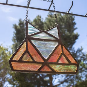 Puede incluir: Un atrapasoles geométrico de vidrieras con una cadena para colgar. El diseño presenta piezas de vidrio naranja, verde y transparente enmarcadas por metal oscuro. El atrapasoles está suspendido sobre un fondo de árboles y cielo azul.