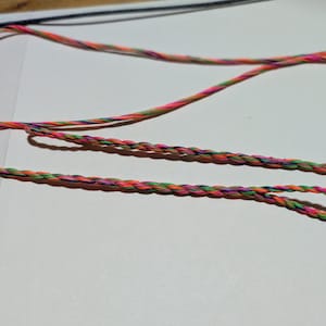May include: Close-up of colorful braided cord with a loop at the end. The cord is a mix of pink, orange, green, and purple. The cord is lying on a white surface.