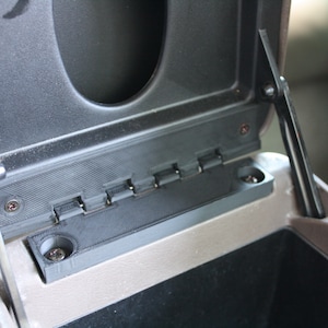 May include: A close-up of a black plastic hinge with multiple metal pins. The hinge is attached to a gray plastic surface with two screws.