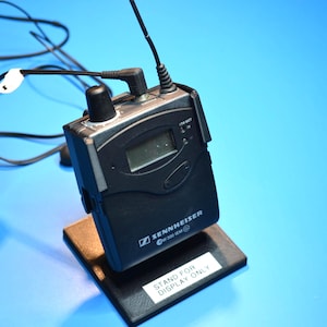May include: Black Sennheiser EW 300 G2 series wireless microphone receiver with a small display screen, antenna, and attached earbuds. The receiver is on a black stand with the text "STAND FOR DISPLAY ONLY".