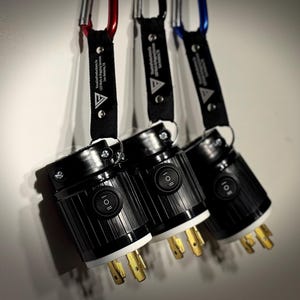 May include: Three black electrical plugs with white and gold prongs are suspended by black straps and carabiners in red, black, and blue. Each plug has an on/off switch and the text "Visual Productions Co. LED Walls & Rigging Services San Antonio, TX".
