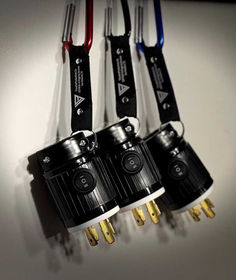 May include: Three black electrical plugs with white and gold prongs are suspended by black straps and carabiners in red, black, and blue. Each plug has an on/off switch and the text "Visual Productions Co. LED Walls & Rigging Services San Antonio, TX".
