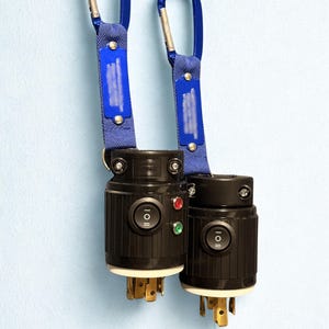 May include: Two black electrical connectors with a switch and indicator lights, attached to blue fabric straps with blue carabiner clips. The connectors have gold-colored prongs and are set against a light blue background.