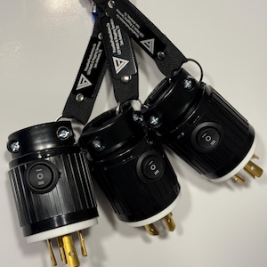 May include: Three black electrical connectors with white and gold prongs are suspended from a carabiner. Each connector has a black switch labeled "I | 0 | I" and is attached to a black strap with white text that reads "Visualize Productions.Co LED Walls & Rigging Services San Antonio, TX".