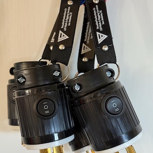 May include: Black electrical connectors with gold prongs and a white base are attached to black straps with the text "VisualizeProductions.Co LED Walls & Rigging Services San Antonio, TX". The connectors have an on/off switch.