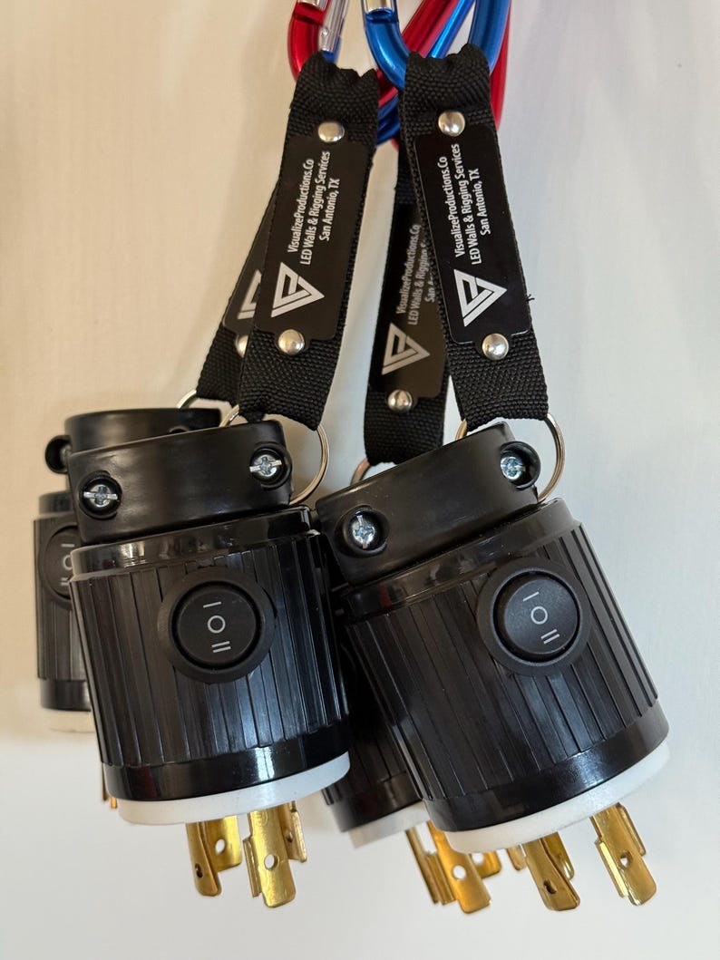 May include: Black electrical connectors with gold prongs and a white base are attached to black straps with the text "VisualizeProductions.Co LED Walls & Rigging Services San Antonio, TX". The connectors have an on/off switch.