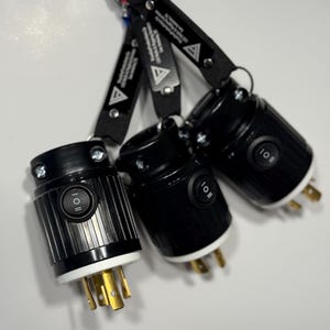 May include: Three black electrical plugs with white and gold prongs are hanging from a carabiner. Each plug has a black on/off switch and is attached to a black strap with white text and a triangle logo.