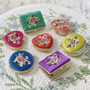 May include: Seven small, ornate, embroidered compacts with mirrors. The compacts are in the shapes of hearts and rectangles. The embroidery features floral designs in shades of pink, red, green, blue, yellow, and purple.
