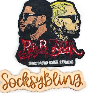 May include: An embroidered patch featuring two figures in profile wearing sunglasses, with the red text R&B TOUR and white text CHRIS BROWN USHER RAYMOND below. Below the patch is a wooden cutout with the word SocksyBling in cursive script.