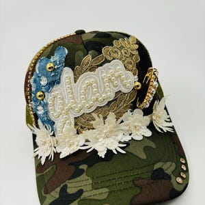 May include: Camouflage baseball cap with the word "Love" in white script, embellished with pearls, gold trim, denim, and white floral accents. The cap has a curved brim and decorative gold studs.