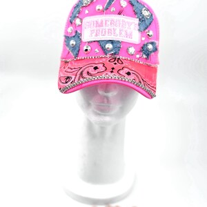 Puede incluir: Una gorra de béisbol rosa brillante con parches de mezclilla desgastados, pedrería transparente dispersa y perlas blancas. Un parche rectangular en la parte delantera muestra el texto SOMEBODY'S PROBLEM. La visera tiene un patrón de cachemira y pedrería.