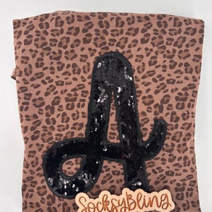 May include: A brown leopard print shirt with a large black sequined letter "A" on the front. A small wooden sign with the text "SocksyBling" is at the bottom. The shirt is folded.