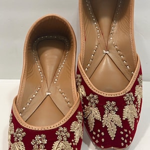 May include: A pair of burgundy velvet shoes with gold embroidered leaf designs. The shoes have a pointed toe and a flat sole.