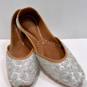 May include: A pair of white flat shoes with silver embroidery. The shoes have a pointed toe and a low heel.