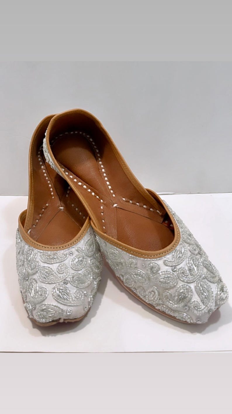 May include: A pair of white flat shoes with silver embroidery. The shoes have a pointed toe and a low heel.