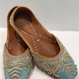 May include: A pair of light blue embroidered flat shoes with gold and silver embellishments. The shoes have a pointed toe and a leather sole.