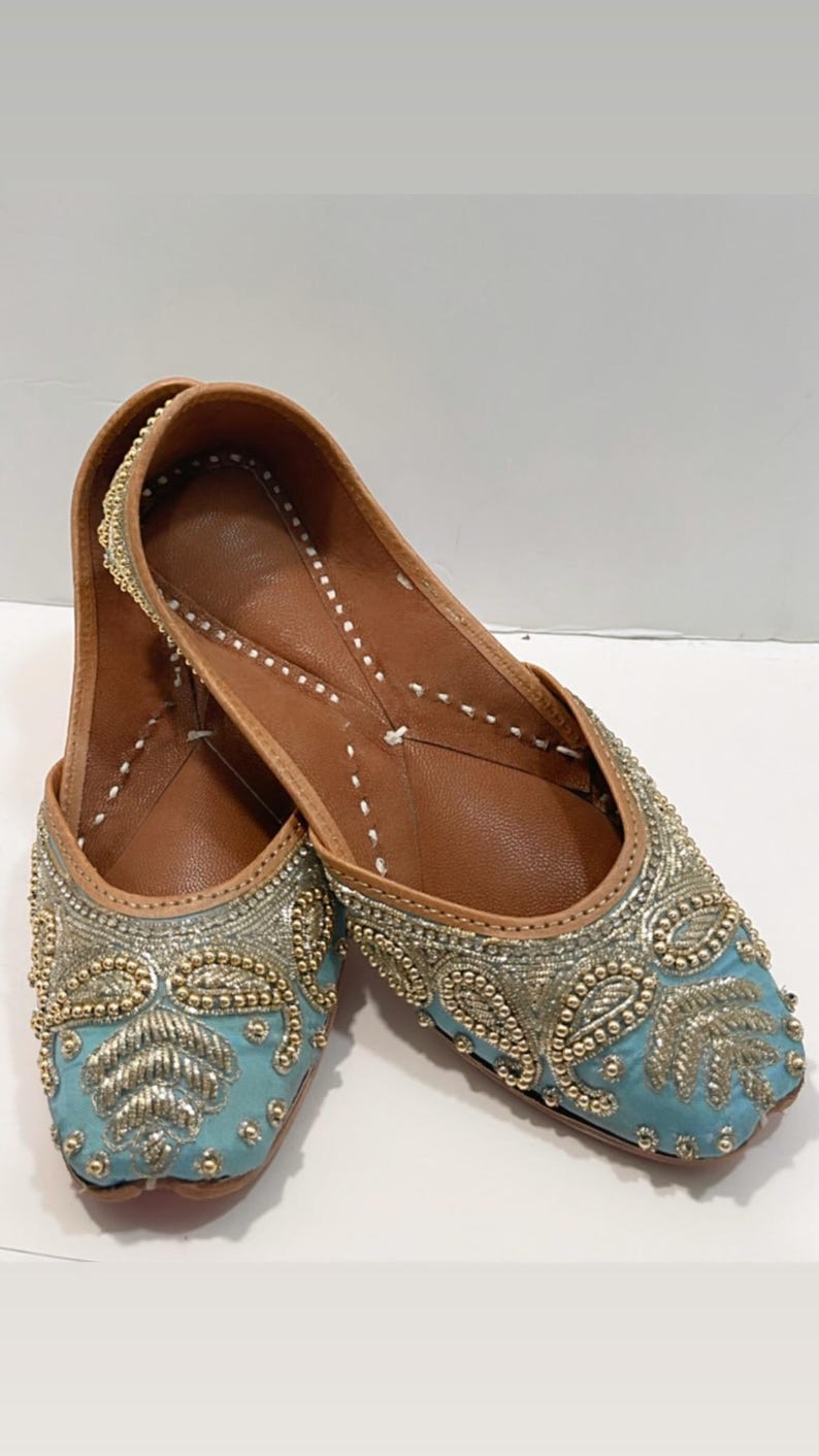May include: A pair of light blue embroidered flat shoes with gold and silver embellishments. The shoes have a pointed toe and a leather sole.