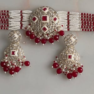 May include: A silver and red beaded choker necklace with a large, ornate pendant and matching earrings. The pendant and earrings are decorated with clear and red gemstones.