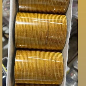 May include: A stack of gold bangles, wrapped in plastic, in a white box. The bangles are shiny and have a slightly textured surface.