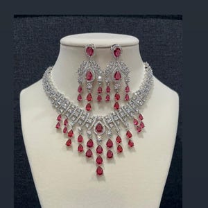 May include: A silver-toned necklace and earring set featuring numerous clear white crystals and multiple teardrop-shaped red stones. The necklace has a multi-layered chain design with dangling red stones, and the earrings match the teardrop motif.