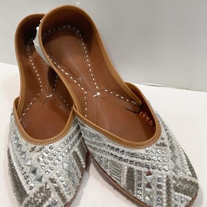 May include: A pair of white flat shoes with silver and white embellishments. The shoes have a pointed toe and a low heel.