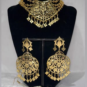 May include: Gold-toned jewelry set featuring a necklace, earrings, and a headpiece. The necklace is a multi-strand design with a floral pendant. The earrings are large, ornate, and dangle. The headpiece is a round pendant with a pearl chain.