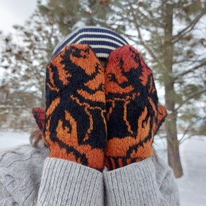 May include: A pair of orange and black knit mittens with a geometric pattern. The mittens are covering the face of a person wearing a striped hat.