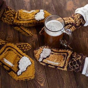 Könnte beinhalten: Ein Bierkrug mit Henkel, gefüllt mit Bier und Schaum, wird von einer Person gehalten, die gestrickte Fäustlinge trägt. Die Fäustlinge sind gelb, braun und weiß, mit einem Bierkrug-Design. Auf einer Holzoberfläche.
