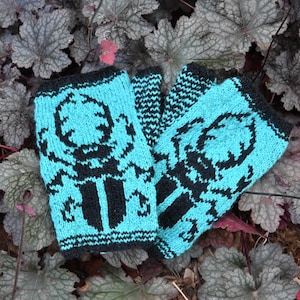 May include: A pair of turquoise fingerless gloves with a black pattern. The gloves are knitted and feature a repeating design. The gloves are resting on a bed of gray and red leaves.