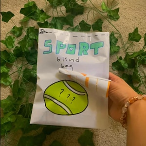 May include: A white paper bag with the word "SPORT" written in blue and green letters. The bag says "blind bag" and has a drawing of a tennis ball with question marks on it.