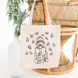 May include: Beige tote bag with a black illustration of a person reading a book, surrounded by butterflies. The bag has a handle and is displayed near a wicker chair and white vases. The words "Fast Delivery" are in a brown box.