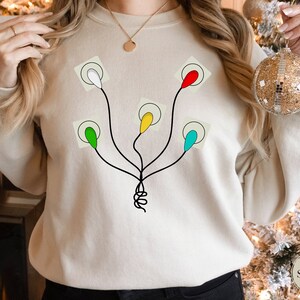 May include: A beige sweatshirt with a graphic of a string of Christmas lights with different colored bulbs. The lights are white, red, yellow, green, and blue.