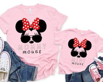 Puede incluir: Dos camisetas rosas con una ilustración en blanco y negro de Minnie Mouse con gafas de sol y un lazo rojo con lunares blancos. La camiseta más grande dice "Mommy Mouse" y la más pequeña dice "Mini Mouse".