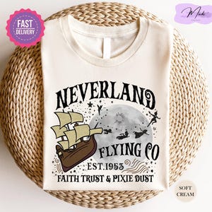 May include: Cream-colored t-shirt featuring a Neverland design. The graphic includes a ship, moon, and silhouettes of Peter Pan and other characters. Text reads "Neverland Flying Co. Est. 1953, Faith Trust & Pixie Dust."