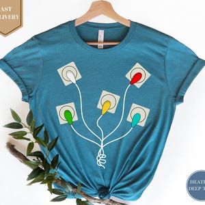 May include: Teal-colored t-shirt featuring a graphic of five colorful light bulbs connected to a central point by white cords. The light bulbs are in green, yellow, red, and teal. The shirt is displayed on a wooden hanger.