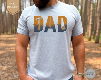 May include: Heather light grey t-shirt with the word "DAD" in large, stylized letters. The letters are designed with tools: a hammer, screwdriver, and saw. The shirt has a crew neck and short sleeves. The background is a blurred forest.
