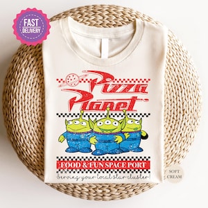 May include: A white t-shirt with a vintage-style graphic of Pizza Planet, a fictional restaurant from the Toy Story franchise. The graphic features three green alien characters from the movie, with the text "Food & Fun Space Port" and "Serving your local star cluster!" below.