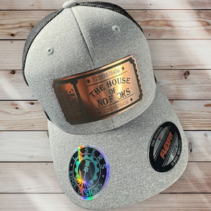 Puede incluir: Una gorra de camionero gris y negra con un parche en forma de boleto de color cobre que dice "The House of No Forks" y una pegatina holográfica con el texto "Laser Edge Designs".