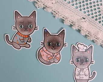 Könnte beinhalten: Drei Katzenaufkleber mit verschiedenen Designs. Die erste Katze trägt einen Pullover mit einem Mond- und Sternenmuster. Die zweite Katze trägt einen rosa Pullover. Die dritte Katze trägt eine Kochmütze und eine weiße Schürze.