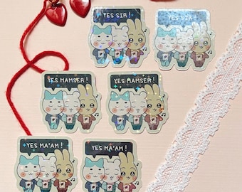 Könnte beinhalten: Eine Sammlung von Aufklebern mit einem Cartoon-Katzen- und Hasenpaar mit den Aufschriften "YES SIR!", "YES MAMSER!" und "YES MA'AM!". Die Aufkleber haben einen holografischen Effekt. Ebenfalls enthalten sind rote herzförmige Ohrringe und eine rote Schnur.
